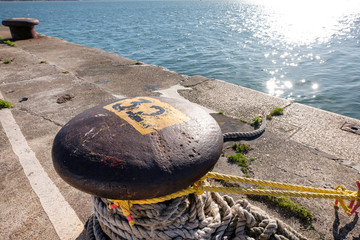 Massiver Poller mit Tauen an einer Kaimauer am Ufer und Sonnenreflexionen im Wasser