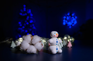 Fototapeta premium A sweet snowman from a pastry mastic on a background of marshmallow trees and a white mug with cocoa. Sweets and a bud of hlok in the dark against the background of a Christmas tree in the lights.