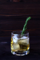 glass of lemonade with pieces of ice. drink in a glass with ice and a sprig of rosemary on a dark wooden background. glass of whiskey or cocktail with ice. vertical