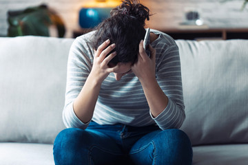 Worried young woman holding her head and mobile phone while sitting on sofa in the living room at home.
