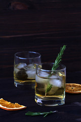 glass of whiskey and ice on wooden table. glass of lemonade with pieces of ice and a sprig of rosemary with slices of orange on a dark background.