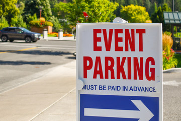 A red, white and blue parking sign on a street sidewalk points towards event parking on a sunny day...