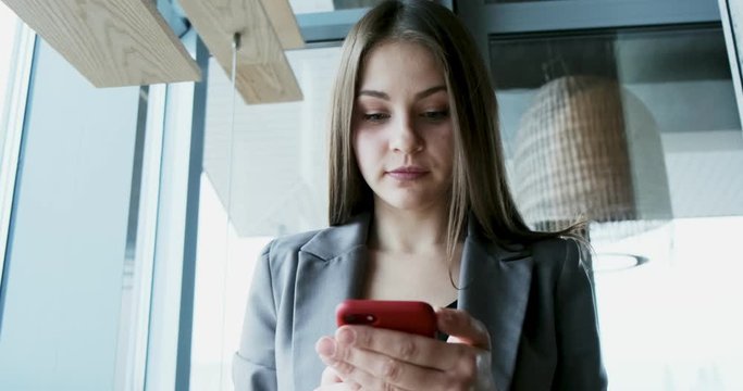 Portrait of young attractive woman looking at smartphone on the office near bif window