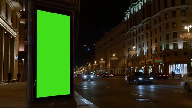 Winter evening. City street. close city light vitrine with empty green screen on roadside against cars driving along night street