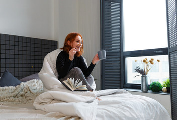 Red hair attractive young woman in bed with cup of tasty coffee and reading book