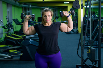 Fototapeta na wymiar An obese woman is working on losing weight. Fat blondes train biceps with a dumbbell in the trainer room. A lot of excess weight due to poor eating habits and lack of self-control.