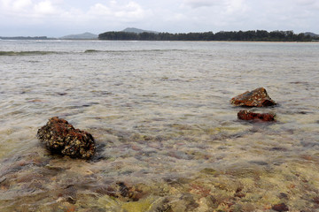 Tropical beach with stones, view from the shallow water to the island with forest and mountains in a ocean. Seascape, concept of vacation on a wild nature