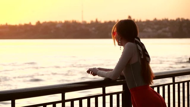Beautiful young woman near railing on waterfront at sunset