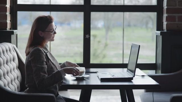 happy girl in glasses communicates with a friend online video communication, using modern gadgets sitting with a laptop at a table in a cafe wifi by the window