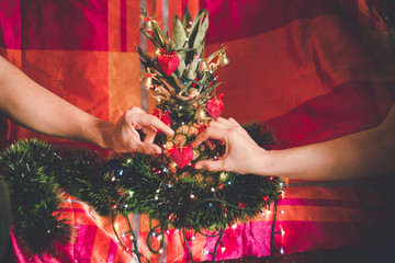 Woman's and man's hands decorating pineapple as alternative Crhistmas tree, with ornaments and Christmas lights on colorful background