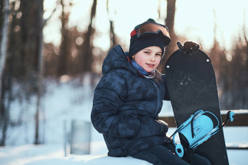 Little kid in protective goggles is posing for photographer with snowboard in winter forest.
