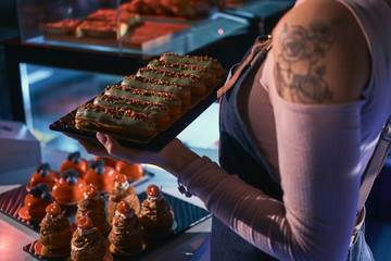 Young lady with tattoo on her shoulder is holding plate with freshly baked eclairs.