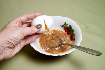 Raw egg on a white plate the table, small spices in it and a spoon in a woman's handon. Cooking eggs for cooking