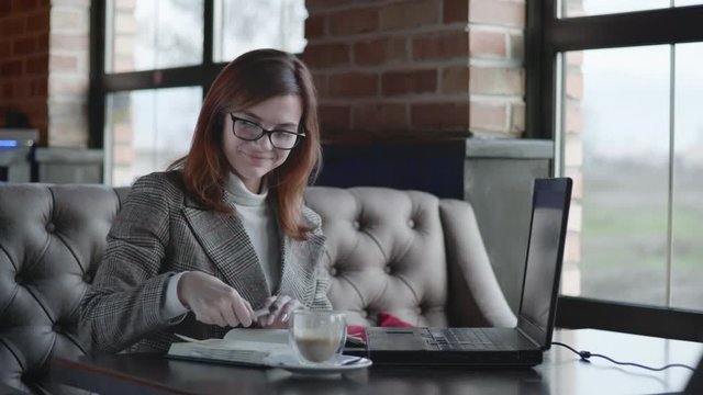 remote work, successful girl in glasses for vision usinesswoman enjoys job online, using laptop computer and writes data to a notebook smiling and looking at the frame while sitting at a table in an