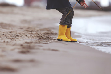 Kid in bright yellow rubber boots is playing at the surf zone of a sandy sea shore - foreground and...