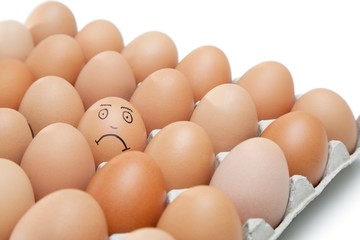 Sad face drawn on an egg surrounded by plain brown eggs in carton against white background