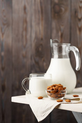 Almond milk in a glass jug and mug, next to nuts in a bowl and a textile napkin on the edge of a white wooden table, wood background.