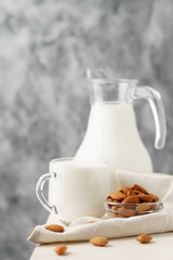Almond milk in a glass jug and mug, next to nuts in a bowl and a textile napkin on the edge of a white wooden table, concrete background.