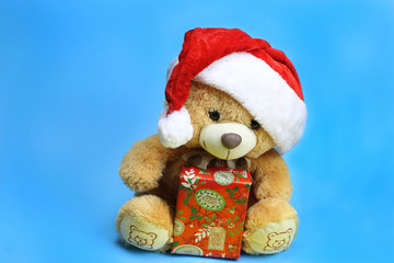 Teddy bear in Santa Claus hat holding a Christmas gift. Blue background