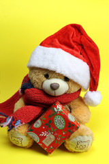 Teddy bear in Santa Claus hat holding a Christmas gift. yellow background
