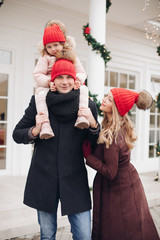 Stock photo of attractive happy Caucasian family of three in red hats in the street. Pregnant mother is looking at her husband with love. Little daughter is sitting on father s neck. They are smiling