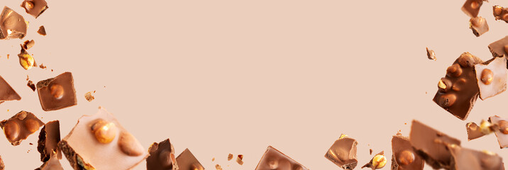 Flying in the air broken bar of milk chocolate with nuts and flakes on pastel pink background. Chocolate pieces levitation concept. Wide banner. frame.