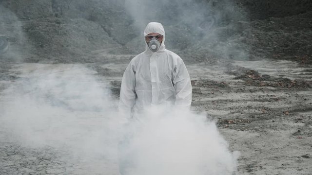 Lab technician in a mask and chemical protective suit, walks on dry ground with a tool box through toxic smoke