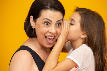 Mom and daughter secretive. kid sharing a secret with mother. Mommy little kid daughter isolated on yellow background studio portrait. Mother's Day love family parenthood childhood concept