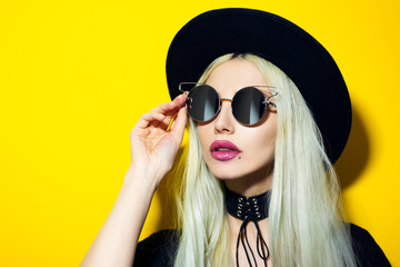 Close-up studio portrait of young blonde girl wearing black sunglasses and hat on yellow background.