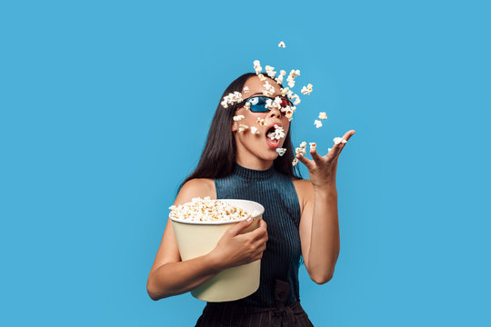 Freestyle. Young woman wearing 3d glasses standing isolated on blue eating popcorn throwing it up trick