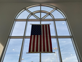 The United States Flag hanging in an interior arched window with a blue sky and white clouds in the background.