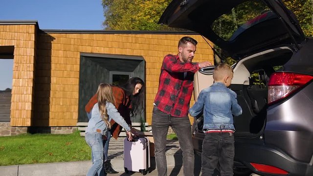 Side view of joyful pleasant family which loading their suitcases into trunk before joint car trip
