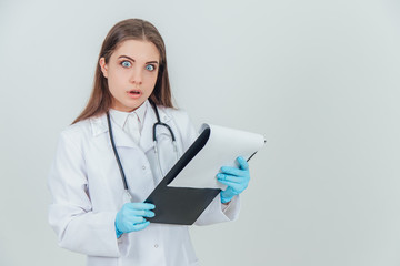 Young female intern standing in medical uniform, holding clipboard, checking data. Surprise and anger in her eyes.