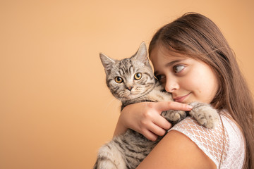 Close up portrait of the beautiful little girl with brunette loose hair the hugging her grey cat, isolated over orange background