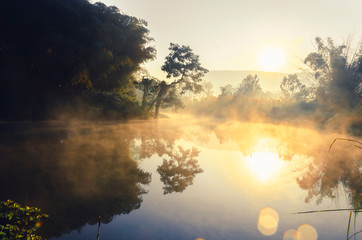 The beautiful landscape of the sunrise, The sun's rays through  at the top of the hill and There is steam above the river, with beautiful Water reflection trees, Chiang Rai Thailand