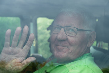 L&auml;chelnder Senior winkt in einem Auto hinter einer Scheibe
