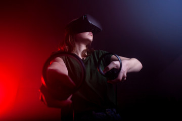 Fototapeta na wymiar girl gamer in modern virtual reality glasses plays a shooter on a dark red-blue background, a woman shoots with game joysticks