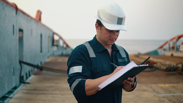 Marine chief officer or chief mate on deck of ship or vessel. He fills up ahts vessel checklist. Ship routine paperwork. He holds VHF walkie-talkie radio in hands.