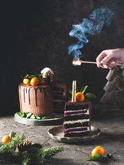 Woman lighting candle on chocolate cake. The cake is decorated with Christmas decor: tangerines, gingerbread house, Christmas tree. Сoncept of sweets and New Year's Eve