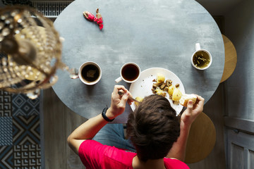 Breakfast at the oval table by the window. Boy eating potatoes and drinking coffee from white ceramic mugs. Top view through a crystal chandelier. Weekend Family Customs Concept