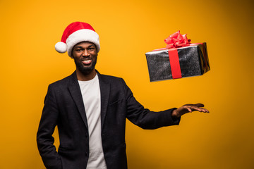 African American man in Santa hat throwing up gift box isolated on yellow background
