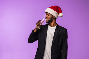 Young african man in santa hat drinking champagne isolated on violet background