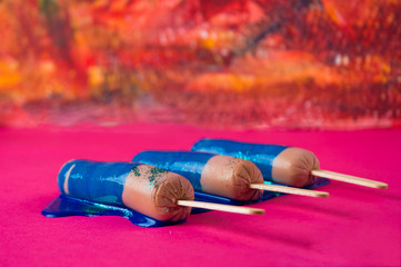 Sausages on sticks in blue sparkle paint. On a pink and orange textured background, top view photo.