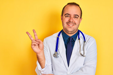 Young doctor man wearing coat and stethoscope standing over isolated yellow background smiling with happy face winking at the camera doing victory sign. Number two.