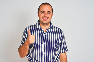 Young man wearing blue striped shirt standing over isolated white background doing happy thumbs up gesture with hand. Approving expression looking at the camera showing success.