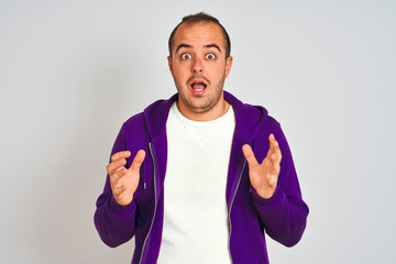 Young man wearing purple sweatshirt standing over isolated white background celebrating surprised and amazed for success with arms raised and open eyes. Winner concept.