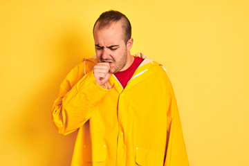 Young man wearing rain coat standing over isolated yellow background feeling unwell and coughing as symptom for cold or bronchitis. Healthcare concept.