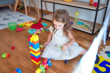 Adorable blonde toddler playing with building blocks around lots of toys at kindergarten