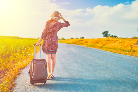 Very cute female tourist in summer dress and hat hitchhiking with suitcase at autumn. Pretty woman walking on the road with Luggage. Hitchhiking concept
