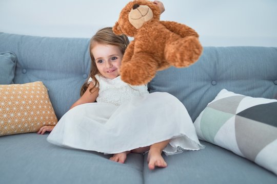 Adorable blonde toddler sitting on the sofa playing with teddy bear at home
